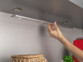 Fixation en cours de la réglette LED sous un meuble de cuisine suspendu à l'aide de deux supports magnétiques adhésifs et d'une main tenant la lampe du bout des doigts pour l'aimanter aux supports