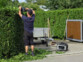 Homme en short et t-shirt bleu marine entrain de tailler une haie d'arbuste avec un taille-haie branché à la batterie nomade et convertisseur solaire HSG-1200 et des outils de jardin placés à côté