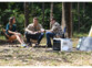 Groupe de 3 amis en forêt assis chacun sur une chaise de camping devant une petite table basse avec à côté une tente jaune montée et la batterie nomade en cours de chargement via une cellule solaire mobile 