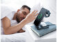Chargement en cours d'une Apple Watch et du boîtier d'une paire d'EarPods sur la station de chargement posé sur un livre sur une table de nuit à côté d'un homme dormant dans un lit aux draps blancs